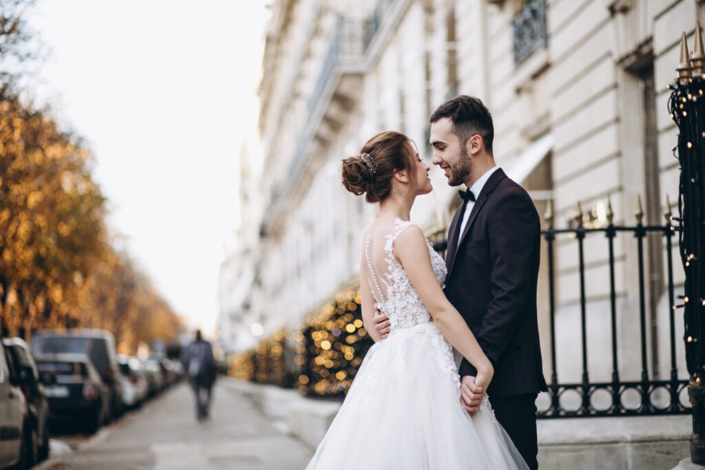 Braut und Bräutigam in eleganter Hochzeitskleidung lächeln sich auf einer städtischen Straße an.