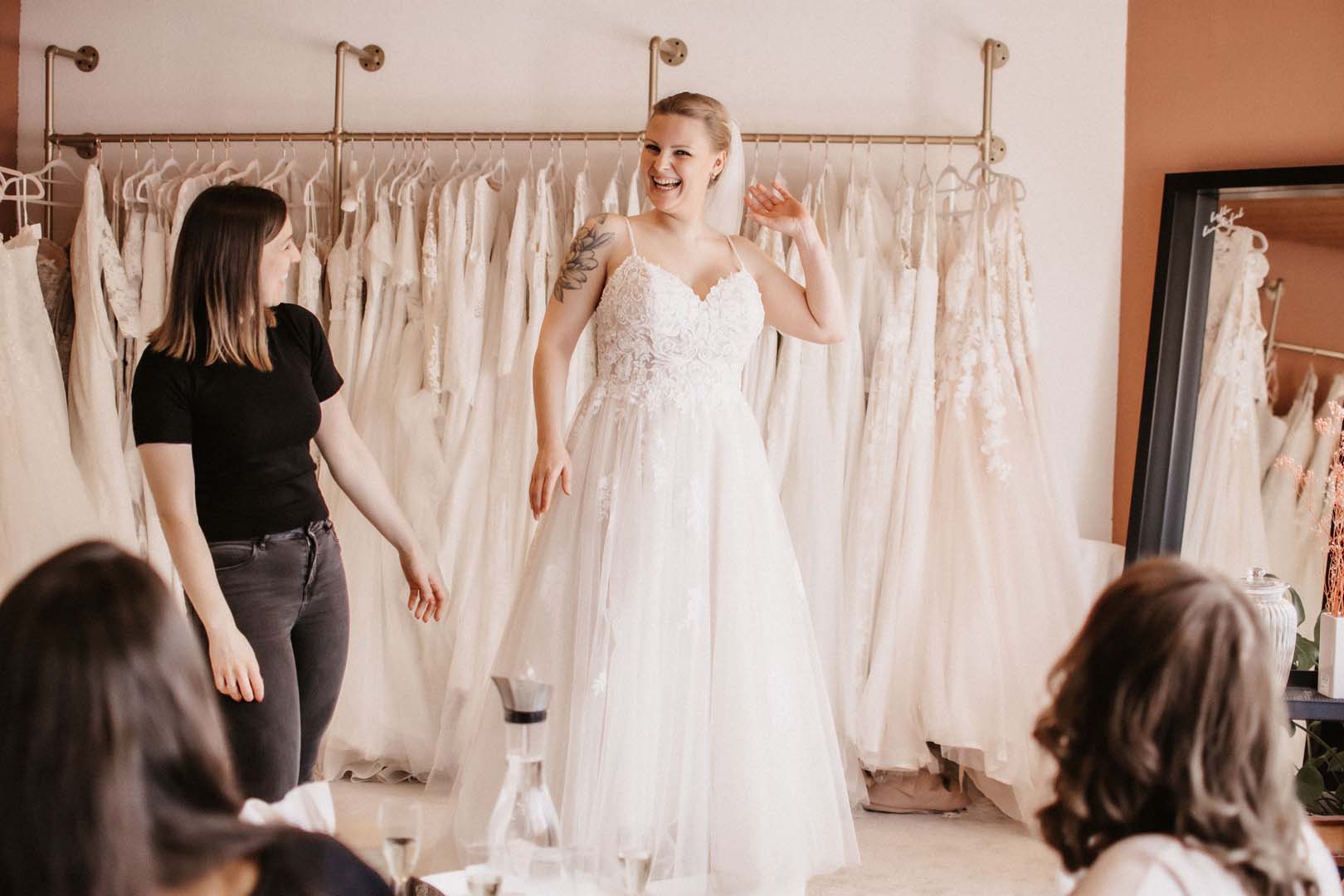 Braut probiert ein Hochzeitskleid in einem Brautladen an, umgeben von Beraterinnen.